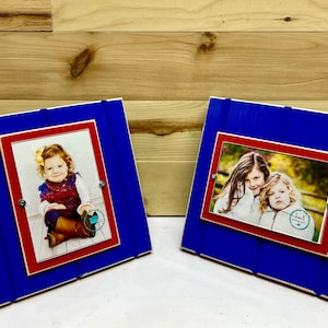 May include: Two blue wooden picture frames with red matting and a white background. The frames are standing upright on a wooden surface. The left frame has a photo of a young girl sitting on a wooden floor. The right frame has a photo of two young girls standing in a field.