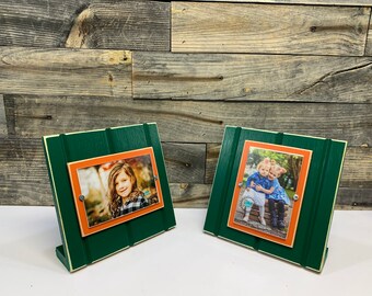 May include: Two green wooden picture frames with orange matting and a white accent line. Each frame holds a photo of children. The frames are standing on a wood surface.