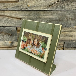May include: A green wooden picture frame with a white border and a photo of two girls in a field. The frame has a stand on the back and is made to look like a miniature house.
