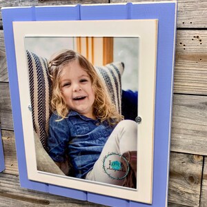 May include: A white picture frame with a light blue border, featuring a photo of a young girl smiling. The photo is taken indoors and shows the girl sitting on a striped couch. The frame has a white mat and a small logo in the bottom right corner that reads "JUNTER photography".