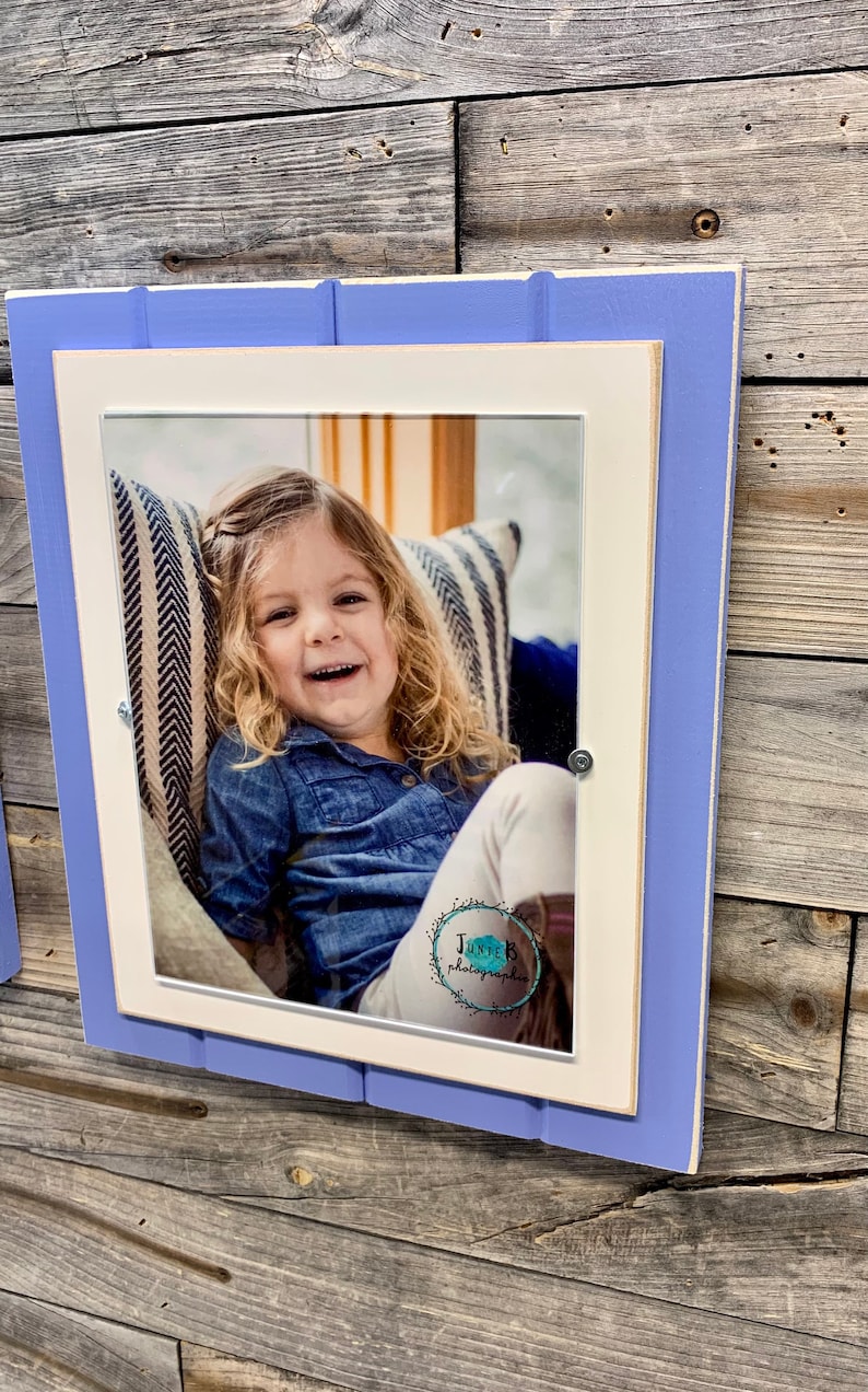 May include: A white picture frame with a light blue border, featuring a photo of a young girl smiling. The photo is taken indoors and shows the girl sitting on a striped couch. The frame has a white mat and a small logo in the bottom right corner that reads "JUNTER photography".