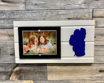 May include: A white wooden frame with a black mat and a photo of two girls in a field. A blue silhouette of a lake is on the right side of the frame.