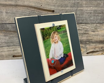 May include: A dark gray wooden picture frame with a white mat and a photo of a young girl sitting on a red cushion. The frame has a built-in stand.