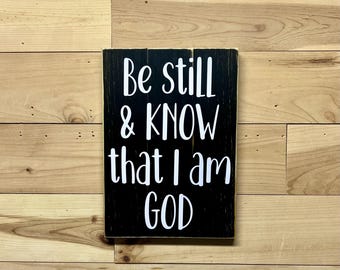 May include: Black wooden sign with white text that reads "Be still & KNOW that I am GOD".