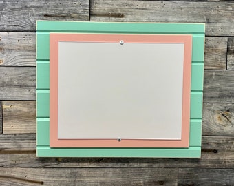May include: A light green framed bulletin board with a light pink inner frame. The board is mounted on a wood panel background.