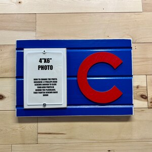 May include: A blue and red wooden sign with a white frame and a red "C" in the center. The sign has a white insert with the text "4"X6" PHOTO" and instructions on how to change the photo.