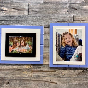May include: Two framed photos on a wood wall. The photo on the left shows two girls sitting on a blanket in a wooded area. The photo on the right shows a girl sitting on a chair with a striped pillow. Both frames are white with a light blue outer border.