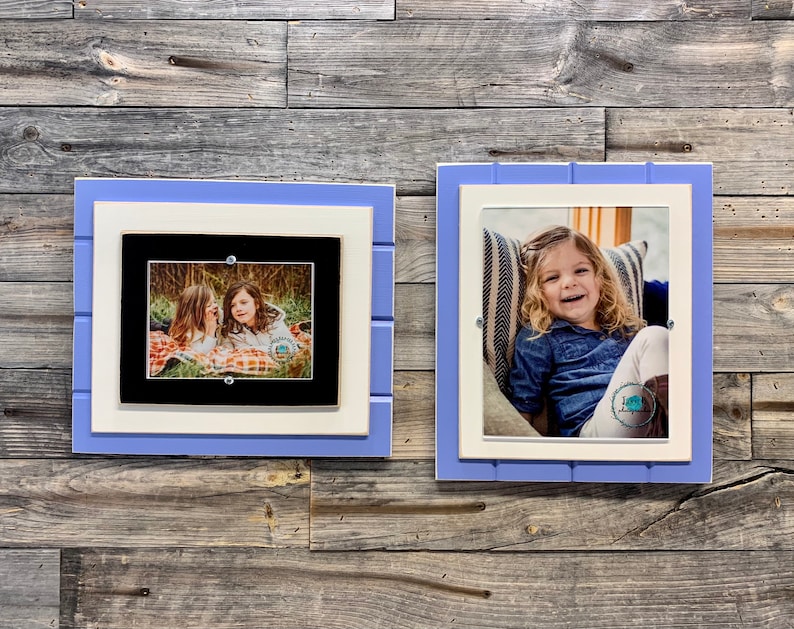 May include: Two framed photos on a wood wall. The photo on the left shows two girls sitting on a blanket in a wooded area. The photo on the right shows a girl sitting on a chair with a striped pillow. Both frames are white with a light blue outer border.