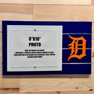May include: A white wooden frame with a blue border and a wood plank background. The frame has a white mat and a clear acrylic insert for an 8x10 inch photo. The frame has a Detroit Tigers logo in orange on the right side. The frame includes instructions on how to change the photo.