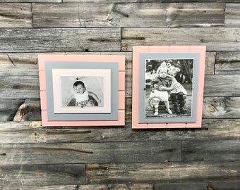 May include: Two framed black and white photographs on a gray wood background. The left frame is pink with a gray inner frame and a photo of a baby. The right frame is pink with a gray inner frame and a photo of two children hugging.