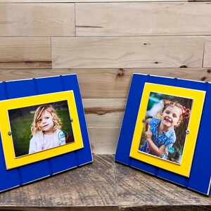 May include: Two blue and yellow wooden picture frames with a white border. Each frame holds a photo of a young girl.