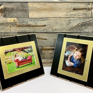 May include: Two black wood picture frames with gold accents and a stand. Each frame has a photo of a young girl inside.
