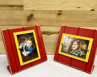 May include: Two red wooden picture frames with yellow trim and a white stand. Each frame holds a photo of a young girl.
