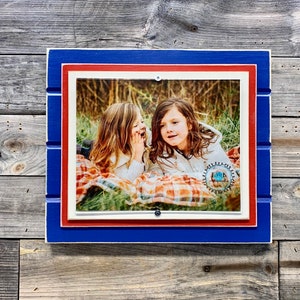 May include: A blue, red, and white wooden picture frame with a photo of two girls in a field. The girls are wearing casual clothing and are smiling. The frame has a rustic look and is perfect for displaying a favorite photo.