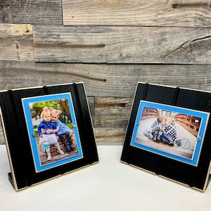 May include: Two black wooden picture frames with blue mounts and a white border. Each frame holds a photograph of two children. The photograph on the left shows two girls sitting on a wooden structure. The photograph on the right shows two children sitting on a wooden bridge.