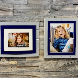 May include: Two framed photos of a young girl, one in a blue and gray frame with a white mat, and the other in a blue and gray frame with a white mat and a striped pillow behind the girl.