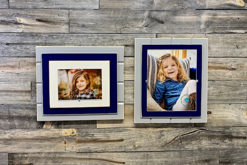 May include: Two framed photos of a young girl, one in a blue and gray frame with a white mat, and the other in a blue and gray frame with a white mat and a striped pillow behind the girl.