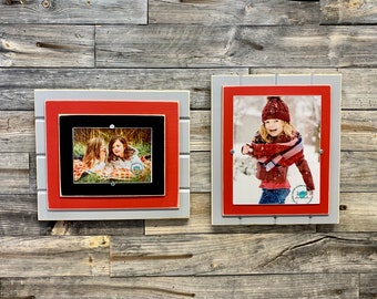 May include: Two framed photos on a wood background. The photo on the left shows two girls sitting on a blanket. The photo on the right shows a girl in a red jacket and hat standing in the snow. Both frames have a red inner frame and a gray outer frame.