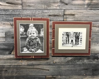 May include: Two rustic wood picture frames with a red and brown finish. The frames are hanging on a gray wood wall. The left frame has a black and white photo of a young girl. The right frame has a black and white photo of a family of three walking.