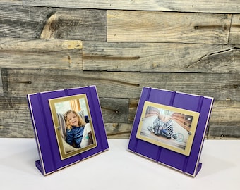 May include: Two purple wooden picture frames with gold trim and a white mat. Each frame holds a photo of a child. The frames are standing on a light wood surface.