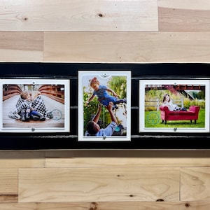 May include: A black wooden picture frame with three white picture frames inside. The frames are arranged horizontally. The left frame shows two children hugging on a bridge. The middle frame shows a man holding a child in the air. The right frame shows a child sitting on a red couch.