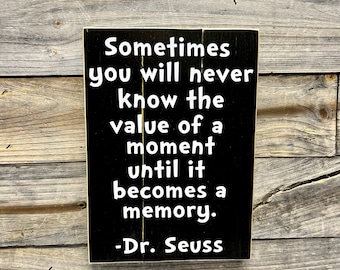 May include: Black wooden sign with white text that reads "Sometimes you will never know the value of a moment until it becomes a memory. -Dr. Seuss"