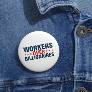 May include: A white button with the text "WORKERS OVER BILLIONAIRES" in navy blue and red. The button is set against a blue denim jacket.