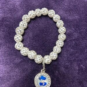 Puede incluir: Un brazalete de cuentas blancas con un dije plateado que presenta una imagen azul y blanca. El dije es redondo y tiene un borde plateado.