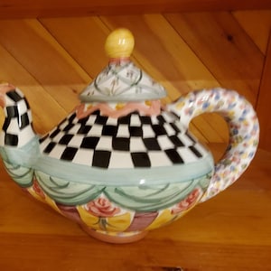 Könnte beinhalten: Eine dekorative Keramik-Teekanne mit einem schwarz-weißen Schachbrettmuster auf dem Körper. Der Griff ist mit bunten floralen Designs verziert. Der Deckel hat einen gelben Knopf und eine gemusterte Oberseite. Die Teekanne steht vor einem Holzgrund.