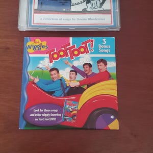 May include: Two children's music CDs. The top CD is titled "Computer Cat" and features a cartoon cat on the cover. The bottom CD is "The Wiggles Toot Toot!" with the band members in a red car. Both CDs are in clear plastic cases.