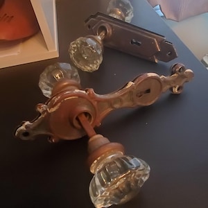 Puede incluir: Juego de herrajes de puerta antiguos que incluye una manija y una placa separada, ambas con un aspecto envejecido y desgastado. La manija tiene un diseño decorativo con pomos de vidrio transparente facetado. Los componentes metálicos muestran desgaste.