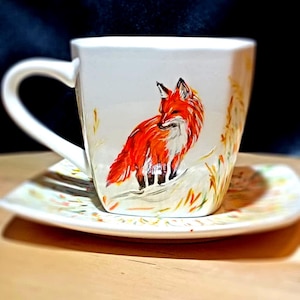 May include: A white ceramic teacup and saucer with a hand-painted design of a red fox standing in a field of yellow and green grass.