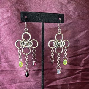 Könnte beinhalten: Silberfarbene Ketten-Ohrringe mit floralem Design. Jeder Ohrring hat eine Ansammlung ineinandergreifender Ringe und hängender Ketten. Die Ketten sind mit farbigen tropfenförmigen Perlen in Grün, Rosa und Schwarz verziert.