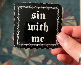 Può includere: Un adesivo quadrato nero con testo bianco che recita "sin with me" in un carattere gotico. L'adesivo è bordato da un disegno di filo spinato. L'adesivo è tenuto in mano.