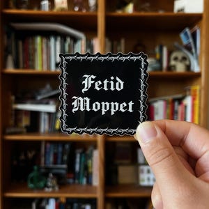 Könnte beinhalten: Ein schwarzer quadratischer Aufkleber mit den Worten "Fetid Moppet" in gotischer Schrift. Der Aufkleber hat einen weißen Stacheldrahtrand. Der Hintergrund ist ein Bücherregal aus Holz, das mit Büchern gefüllt ist.