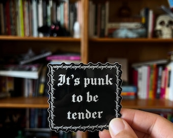 Può includere: Un adesivo quadrato nero con bordi in filo spinato bianco e la scritta "It's punk to be tender" in un carattere gotico. L'adesivo è tenuto davanti a una libreria piena di libri.