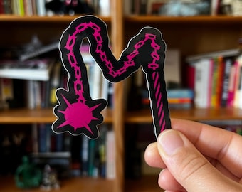 May include: A black sticker in the shape of a flail, with a pink chain and spiked ball design. The handle has pink stripes. The sticker is held in front of a bookshelf.