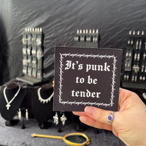 May include: A black square patch with white text that reads "It's punk to be tender" surrounded by a barbed wire design. The image also shows various silver necklaces, earrings, and a small gold-framed mirror on a display table.