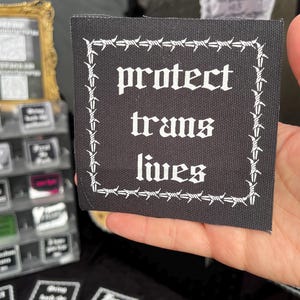May include: A black square patch with white text that reads "protect trans lives." The patch is bordered by a white barbed wire design. The patch is being held up by a hand.