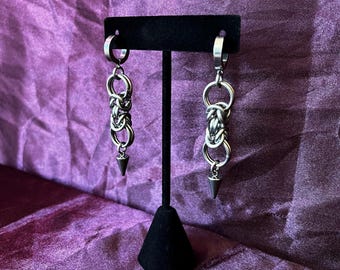 May include: A pair of silver earrings. Each earring has a hoop at the top, linked to interwoven metal links, and a small, pointed cone at the bottom. The earrings are displayed on a black stand against a purple backdrop.