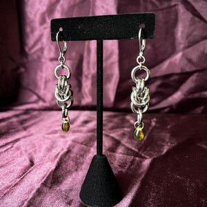 May include: A pair of silver-toned earrings with a chainmail design, each featuring a small, olive-green teardrop-shaped bead. The earrings are displayed on a black stand against a deep purple background.