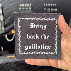 May include: Square black patch with white barbed wire border and text. The text reads "Bring back the guillotine" in a gothic font. The patch is held up by a hand.