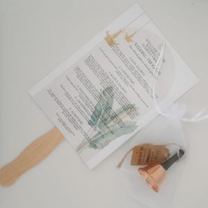 Puede incluir: Un abanico de programa de boda con un diseño de hojas tropicales. El abanico está hecho de papel y tiene un mango de madera. El programa incluye el horario de la ceremonia de boda, con eventos como "Jaan Aagman", "Ganesh Puja" y "Kanya Aagman".