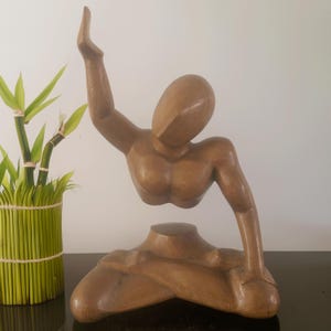 May include: A wooden sculpture of a person in a seated position with one arm raised. The sculpture is made of light brown wood and has a smooth finish.
