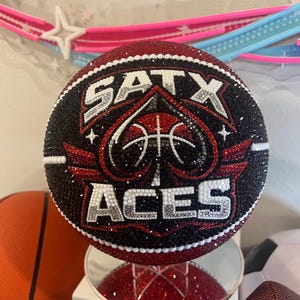 May include: A bedazzled basketball featuring the text "SATX ACES" in white and silver rhinestones. The basketball has a black and red design with a basketball and ace of spades logo. The ball is displayed on a mirrored surface.