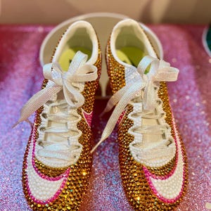 Op de afbeelding: Een paar versierde sportschoenen. De schoenen zijn bedekt met gouden en heldere strass-steentjes, met witte accenten en roze randen. Witte veters en een wit swoosh-ontwerp zijn zichtbaar. De binnenkant is wit met een groene binnenzool.