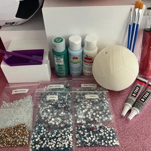 May include: A DIY craft kit featuring a white volleyball, three bottles of acrylic paint in green, light blue, and white, two paintbrushes, a red ball pump, two tubes of B-7000 adhesive, a purple tray, and several bags of assorted rhinestones.