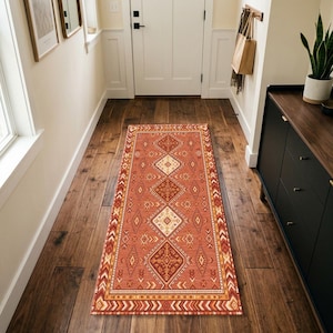 May include: A rectangular runner rug featuring a terracotta orange base with a repeating geometric pattern in cream, dark brown, and mustard yellow. The design includes diamond shapes and chevron borders, centered on a dark wood floor in a hallway.