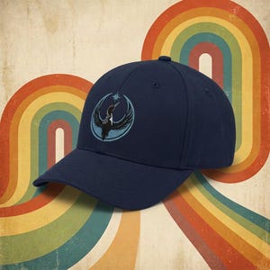 May include: Navy blue baseball cap with a curved brim. The cap features an embroidered emblem of a bird within a circular design, with a star above it. The background has a retro rainbow design.