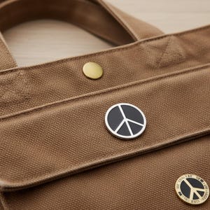 May include: A close-up of a brown canvas tote bag featuring a gold button and a black and silver peace sign pin. The bag has a handle and a pocket with another peace sign pin.